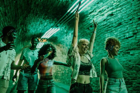 Young people raising arms and dancing at music festival in green lit tunnel