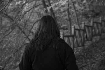 Woman walking lonely on serene forest path with wooden fence in black and white. Melancholic autumn stroll. Deep in thought. Back view