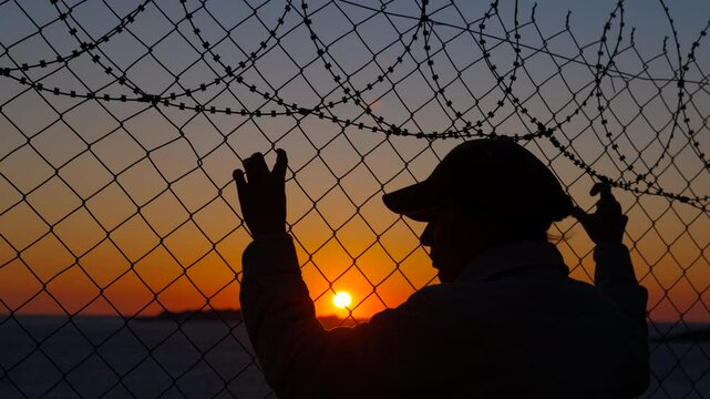Prisoner looking at sunset through barbed wire. Prisoner silhouette holding barbed wire, gazing longingly through fence at sunset, symbolizing hope and freedom amid confinement