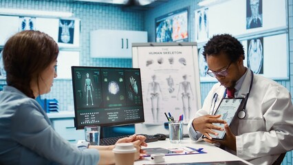 Fototapeta premium African american specialist showing the human skeleton to explain diagnosis to patient, x ray imaging scan interpretation. Medical expert reviewing lab test results in cabinet. Camera B.