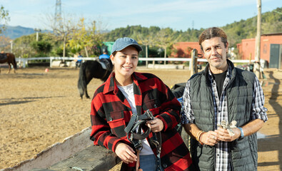 Portrait of two positive male female grooms in plaid shirts posing on yard of horse riding club on...