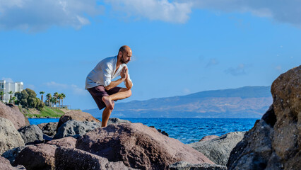 A middle-aged charismatic man, wearing a white shirt, is relaxing and enjoying the sun by doing yoga poses on the rocks by the seaside on a sunny summer day