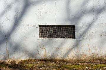 Rectangular crawl space vent, ventilation opening with a metal mesh cover. White aging stucco wall, surrounded by cracks and weathering
