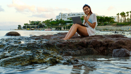 A young red-haired business woman, wearing a beach dress, is sitting on the rocks by the seaside on a sunny summer day, joyfully working remotely on her laptop while on vacation
