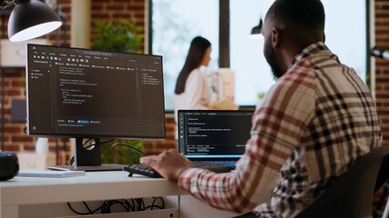 African american IT professional types programming code on dual monitors in home office, working on software development and internet security. Ideal setup for remote work and productivity. Camera B.