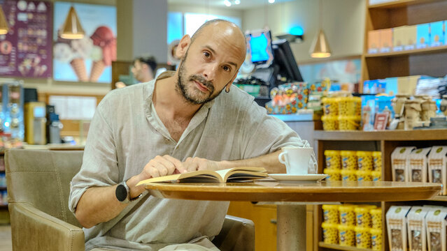 A charismatic middle-aged man wearing a shirt is sitting in a lovely café on a summer evening, enjoying reading a book - Powered by Adobe