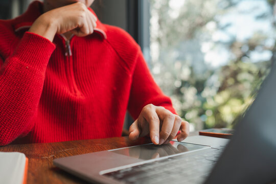 Individual in a red sweater using a laptop indoors, seated at a wooden table with natural light streaming in.