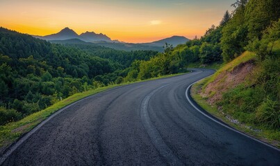 Naklejka premium A quiet road weaves through a forest, the distant mountains looming as the sunset paints the sky with tranquil hues of orange and pink.