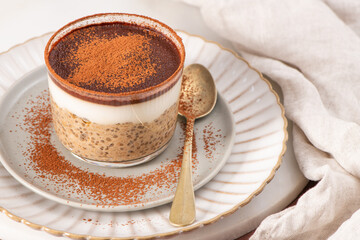 Tiramisu chia pudding in a glass, topped with cocoa powder, on a white plate with a gold spoon, and a beige cloth napkin beside it