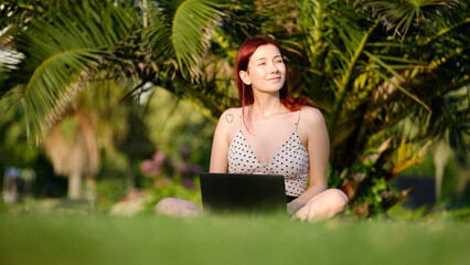 A beautiful, young, red-haired businesswoman sitting on the grass in a tree-filled park, happily...