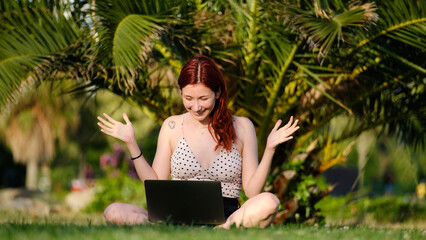 A beautiful, young, red-haired businesswoman sitting on the grass in a tree-filled park, happily...