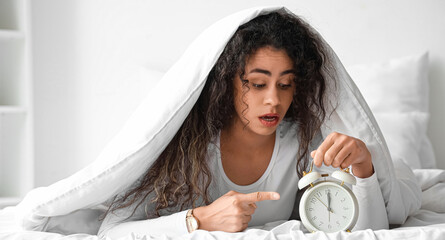 Overslept young African-American woman with alarm clock lying under blanket in bedroom
