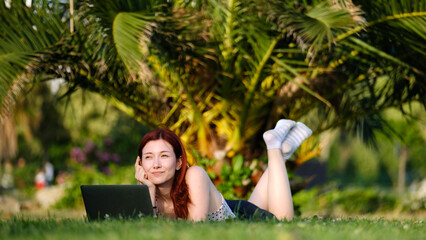 A beautiful, young, red-haired businesswoman sitting on the grass in a tree-filled park, happily...
