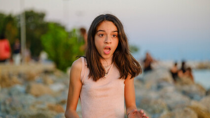 A dark-skinned teenage girl sitting on the rocks by the seaside on a sunny summer day, showing a surprised expression
