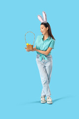 Young Asian woman in bunny ears with basket of Easter eggs on blue background