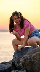 A slightly overweight teenage girl posing cutely while sitting on rocks by the seaside on a sunny summer day