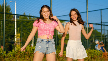 Two teenage girls friends, one slightly overweight and the other with dark skin, dancing and having fun together by the seaside near basketball courts on a sunny summer day