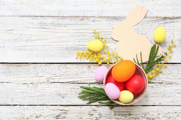 Basket with painted Easter eggs decorated with mimosa flowers on white wooden background