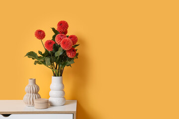 Vase with beautiful orange dahlia flowers on chest of drawers against yellow background