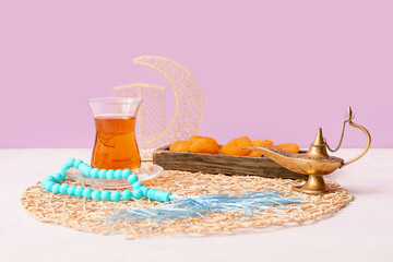 Aladdin lamp, prayer beads, glass cup of tea and dried apricots on white table against lilac background. Ramadan celebration