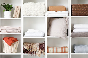 Shelving unit with clean bed sheets, baskets and houseplant near white wall in room