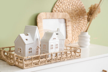 Wicker tray with house shaped candle holders on chest of drawers near green wall in room, closeup