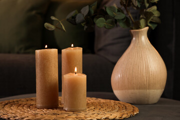 Burning candles and vase with eucalyptus on pouf in dark living room, closeup