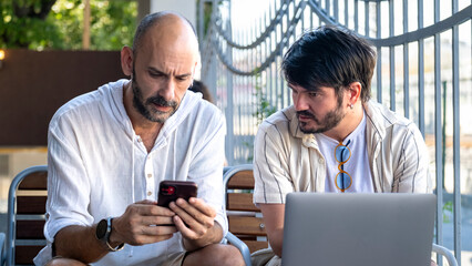 Two middle-aged male professionals are happily working on their laptops in the garden of a café on...