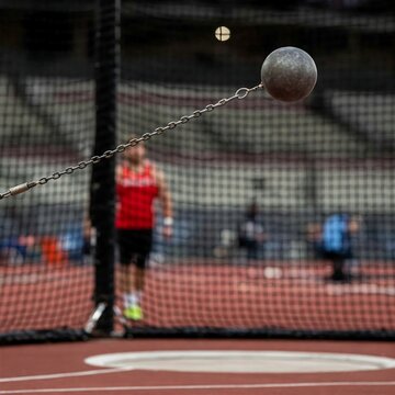  slightly blurred action shot captures a dark, spherical hammer throw weight in mid-flight during a track and field competition