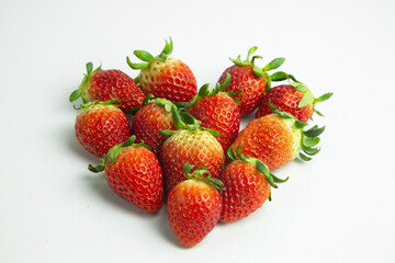 strawberries are arranged in a heart shape on a white background. The berries are red with green stems, some showing a transition to lighter, unripe areas. The image is a top-down view