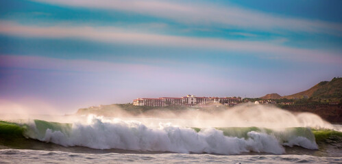 Large wave crashes down with force along the Southern California shoreline with landscape and business in the background