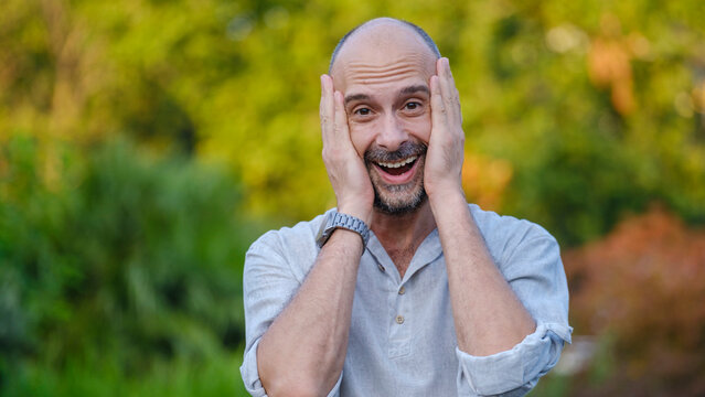 A middle-aged handsome businessman looking at the camera with a surprised expression in a tree-lined park on a summer day - Powered by Adobe