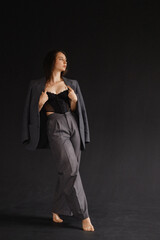 Young woman in a gray jacket thrown over shoulder, high-waisted trousers and a black corset. Caucasian model poses on a dark background. Concept of modern fashion and self-expression.