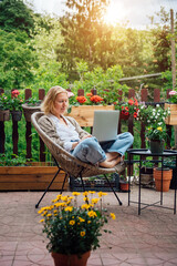 Young adult Caucasian blonde woman sitting on cottage terrace using laptop, online work in the...