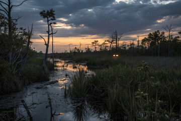 Obraz premium A tranquil wetland at sunset, with the sun shining through the clouds, casting a warm glow over the scene. A vehicle's headlights are visible in the distance, adding a sense of mystery and depth.