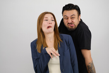 A young businesswoman in a suit and a middle-aged man wearing a black t-shirt stand in front of a white background in a studio environment, making funny facial expressions and looking at the camera