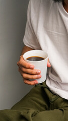 Close-up of a man sitting holding a glass of coffee. The man is wearing a white shirt and green pants.