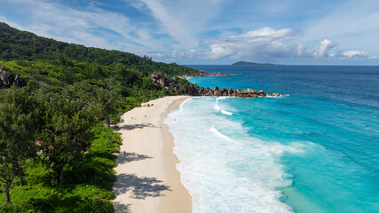 Pristine sandy beach lined with granite rocks and turquoise waters, bordered by lush greenery. La Digue, Seychelles. Grand Anse Beach.
