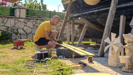 A middle-aged man measuring with a tape measure to make repairs with lumber next to his boat in the garden of his house on a summer day