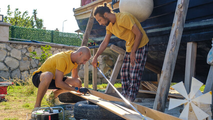 Two middle-aged men measure with a tape measure next to their boat in the garden of their house on...