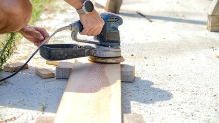 A middle-aged man sanding timber with a sander in the garden of his house on a summer day
