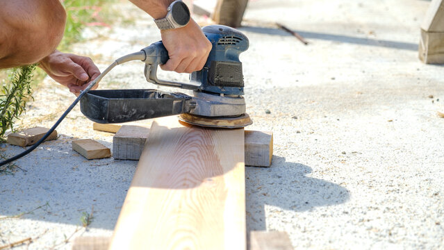 A middle-aged man sanding timber with a sander in the garden of his house on a summer day
