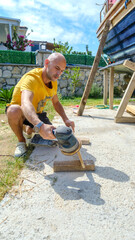 A middle-aged man sanding timber with a sander in the garden of his house on a summer day