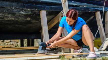 A young woman is sanding timber with a sander in the garden of her home on a summer day.