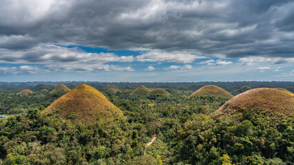 Obraz premium Rows of rounded conical hills covered with brownish grass stretch to the horizon. Thickets of tropical vegetation in the valley. Blue sky, clouds. Philippines. Bohol. Chocolate Hills Natural Monument