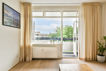 A spacious living room featuring large windows that allow natural light to flood in, paired with soft curtains and a touch of greenery, creating a welcoming atmosphere.