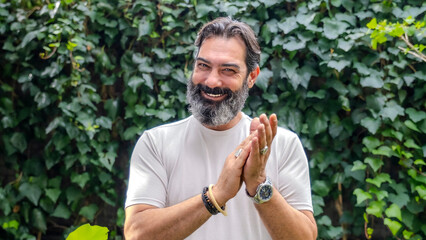 A middle-aged man with a fair complexion and a beard is smiling, looking at the camera, and making a clapping gesture in a close-up shot in a wooded area