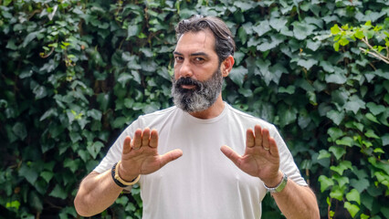 A middle-aged,  man with dark hair and a beard, looking at the camera and making a "stop" gesture with his hand in a wooded area