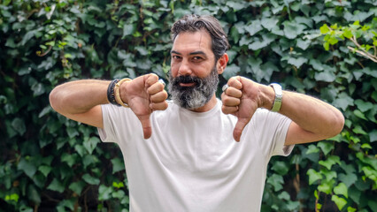 A middle-aged, fair-skinned man with a beard, in an outdoor wooded area, looking at the camera and making a "no" gesture by pointing his thumb downward