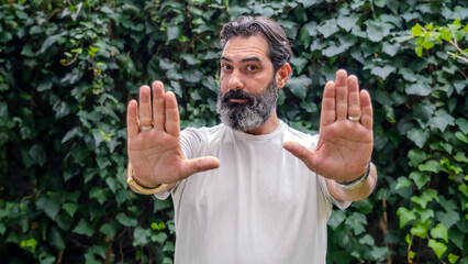 A middle-aged,  man with dark hair and a beard, looking at the camera and making a "stop" gesture with his hand in a wooded area